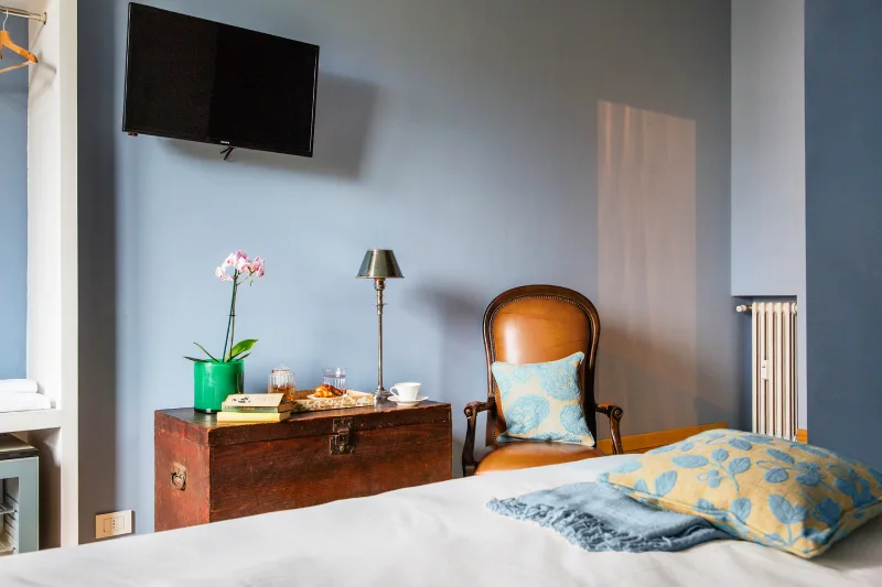 Room in Rome Monti district with blue walls, wooden trunk, vintage armchair, wall TV and light blue cushions on the bed - Salotto Monti, BZAR hotels in Rome