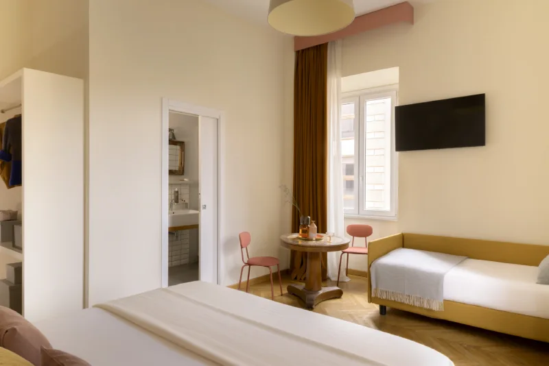 Triple room in Rome Monti area with double bed, sofa bed, table with pink chairs, bright window and bathroom entrance view - Salotto Monti, BZAR hotels in Rome