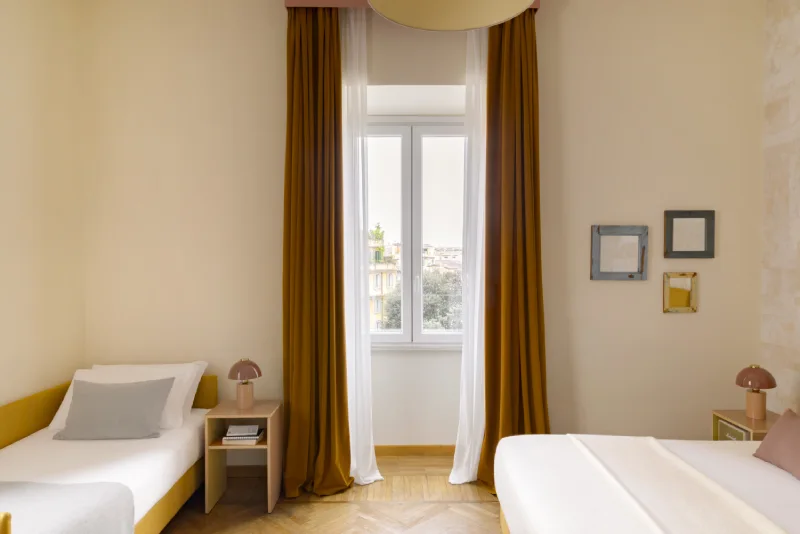 Triple room in Rome Monti district with double and single bed, ochre curtains, bedside tables with pink lamps and bright central window - Salotto Monti, BZAR hotels in Rome
